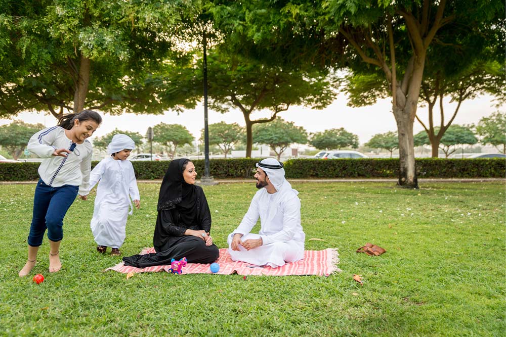 A smiling family with young children having a picnic on a lush green lawn in a family-friendly community in Ajman, showcasing the city's lifestyle.